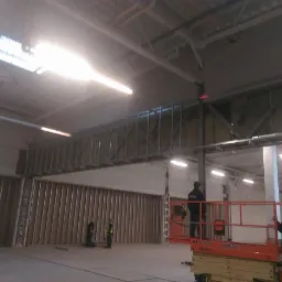 Montaż metalowej konstrukcji ścian działowych w hali, widoczni pracownicy na podnośniku koszowym i przy ścianie, surowy stan wykończenia.