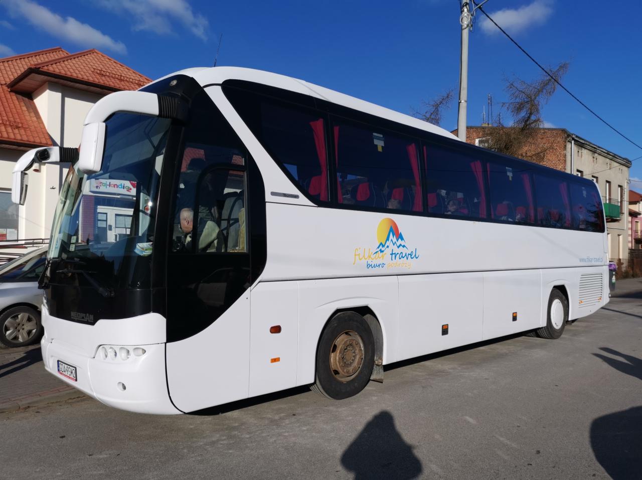 Biały autokar z logo 'filkar travel biuro podróży' na tle miejskiej ulicy i błękitnego nieba.
