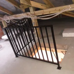 Czarna metalowa balustrada zabezpieczająca otwór w stropie na poddaszu, widok na surowy stan wykończenia, instalacje elektryczne pod sufitem.