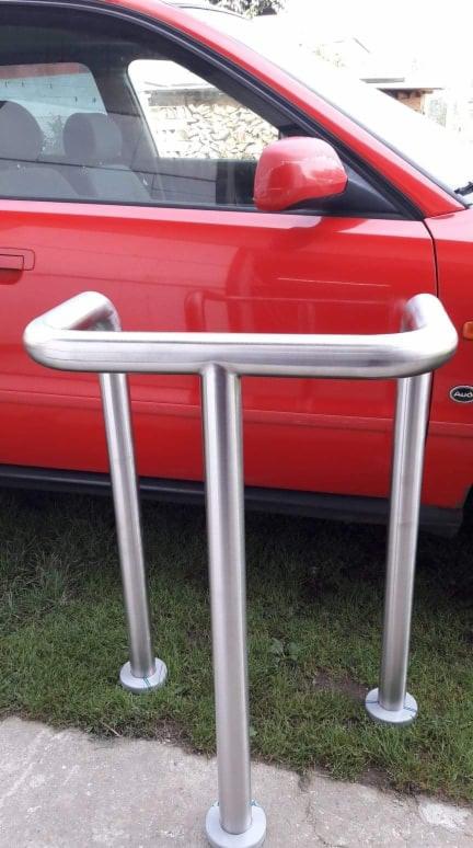 Nierdzewna poręcz zewnętrzna z trzema słupkami na trawniku, z czerwonym samochodem w tle.