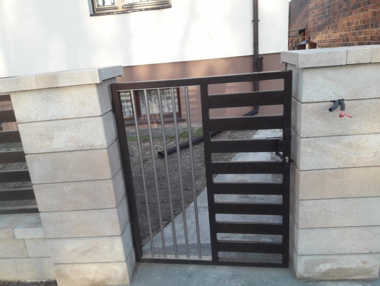 Brązowa furtka z poziomymi szczeblami i pionowymi prętami, zamontowana między słupkami z szarego betonu łupanego.