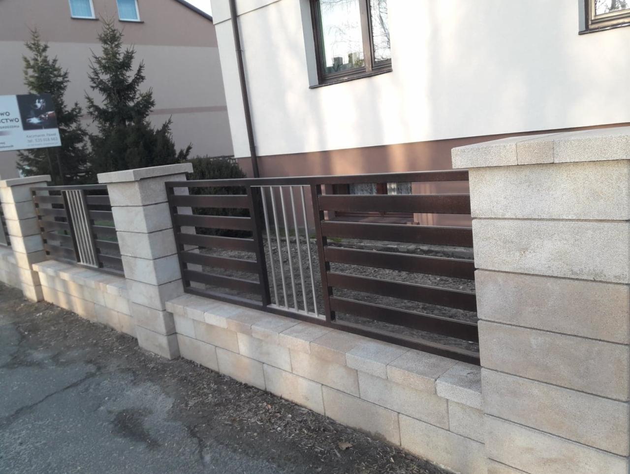 Nowoczesne ogrodzenie z poziomych, brązowych profili metalowych i słupków z szarego betonu modułowego, z fragmentem furtki z pionowych prętów, na tle domu.