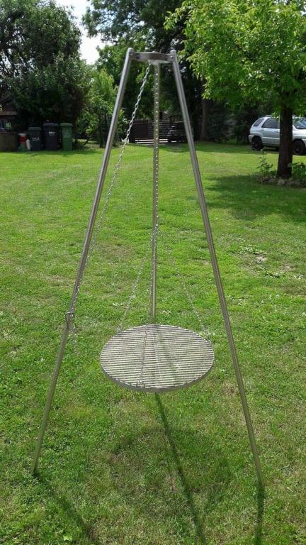 Metalowy trójnóg z rusztem wiszącym na łańcuchach, ustawiony na trawniku w ogrodzie.