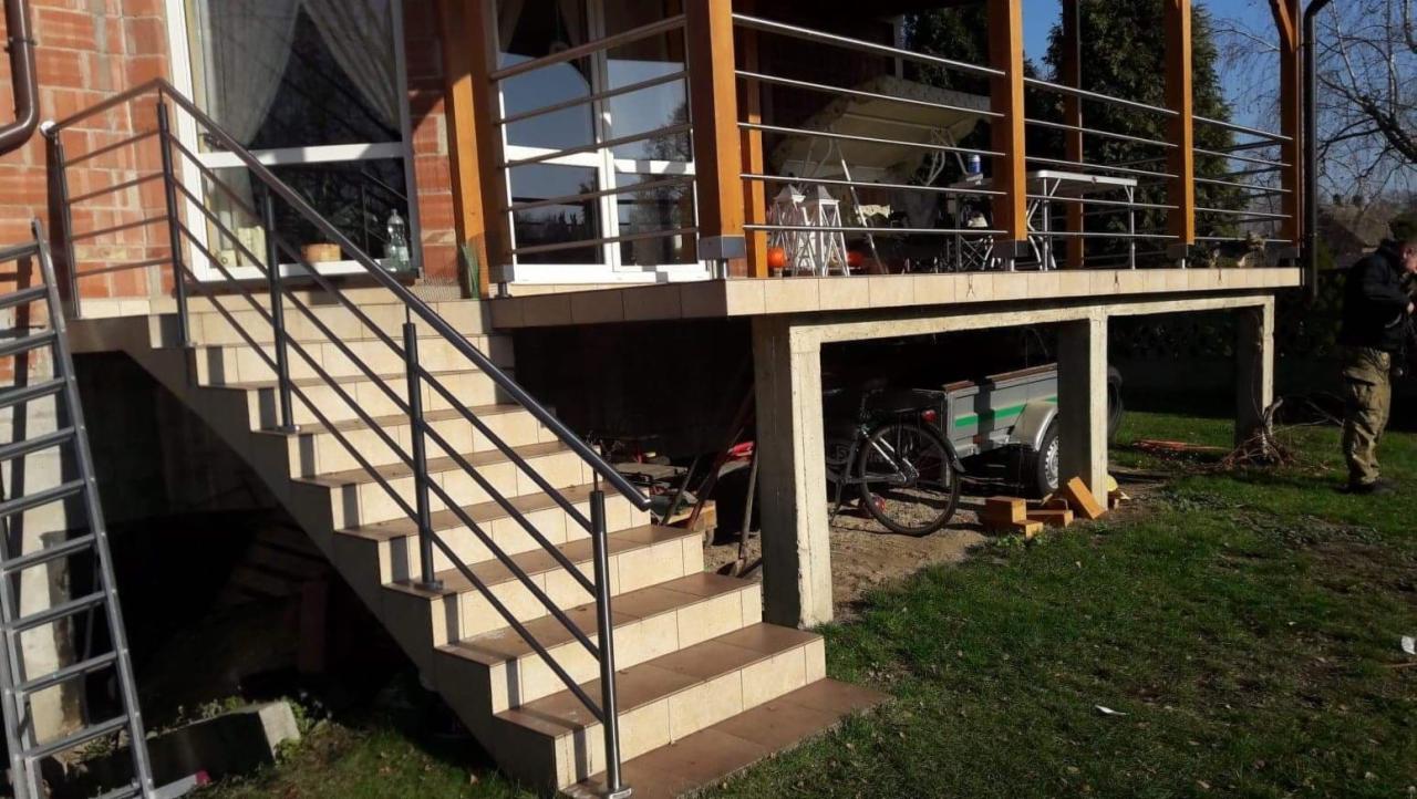 Zewnętrzne schody z beżowych płytek z czarną metalową balustradą i poręczą, prowadzące na taras z drewnianą konstrukcją, w tle widoczny rower i przyczepka.