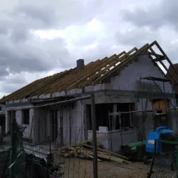 Budowa domu jednorodzinnego w stanie surowym otwartym: wzniesione mury z betonu kom&oacute;rkowego i częściowo zamontowana drewniana konstrukcja dachu pod pochmurnym niebem.