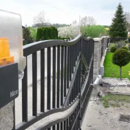 Automatyczna brama wjazdowa z metalowymi sztachetami, wyposażona w lampę ostrzegawczą marki Nice, na tle zadbanego ogrodu z kulistymi krzewami i kwitnącymi tulipanami.