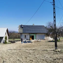 Dwóch dekarzy montuje ciemnoszarą blachodachówkę na drewnianej konstrukcji dachu domu jednorodzinnego w słoneczny dzień. Na ziemi leżą arkusze blachy, a w tle widać krajobraz.
