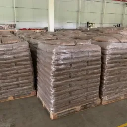 Magazyn wypełniony paletami z workami brązowego granulatu, owiniętymi folią, ustawionymi w rzędach, widoczne elementy konstrukcji hali.