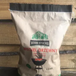 Brązowa papierowa torba z węglem drzewnym 'Wood Stocks' 2,5 kg, na tle drewnianej ściany. Na torbie nadruk z drzewami i grillem.