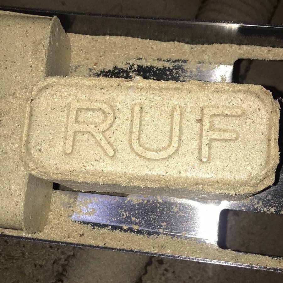 Brykiet RUF z widoczną strukturą i napisem na metalowej powierzchni, otoczony drobnymi trocinami.