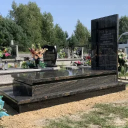 Czarny, polerowany pomnik z granitu z inskrypcjami, otoczony piaskiem i kwiatami na cmentarzu, w tle inne nagrobki i biały samochód dostawczy.