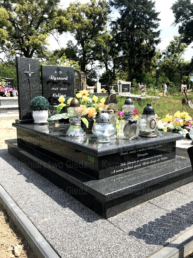 Czarny, dwupoziomowy pomnik z inskrypcjami i dekoracjami kwiatowymi oraz lampionami na cmentarzu, z widocznym fragmentem ścieżki z szarej kostki brukowej.