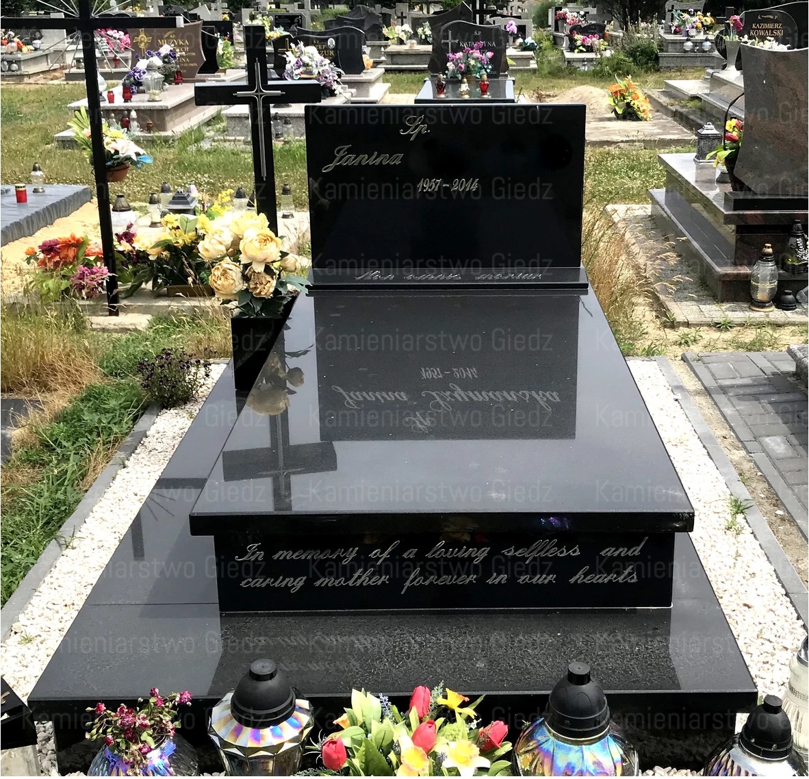 Czarny, polerowany pomnik z wygrawerowanym imieniem, nazwiskiem i datami, otoczony kwiatami i zniczami na cmentarzu.