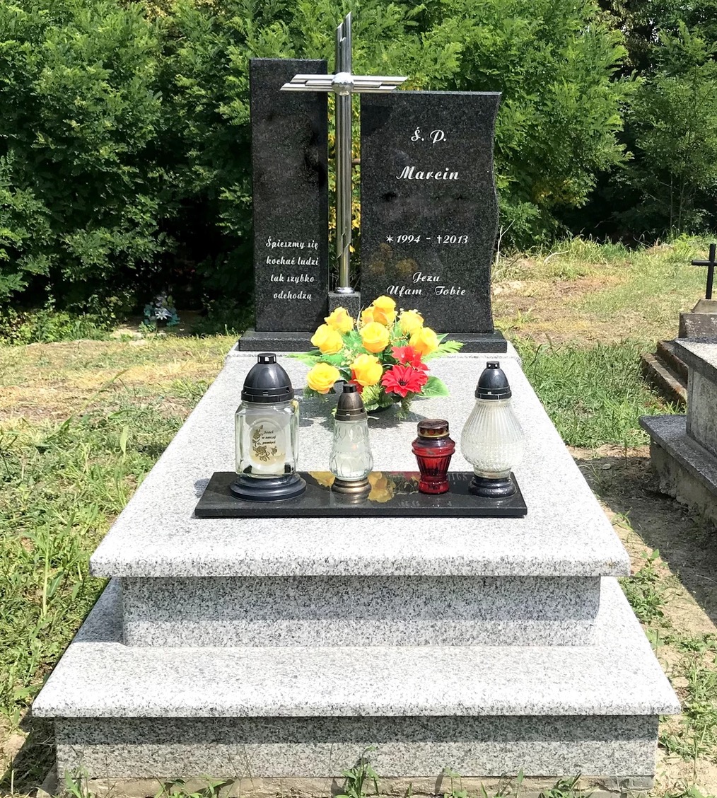 Granitowy pomnik z krzyżem, świeżymi kwiatami i zniczami na dwupoziomowej podstawie, na tle zieleni cmentarza.