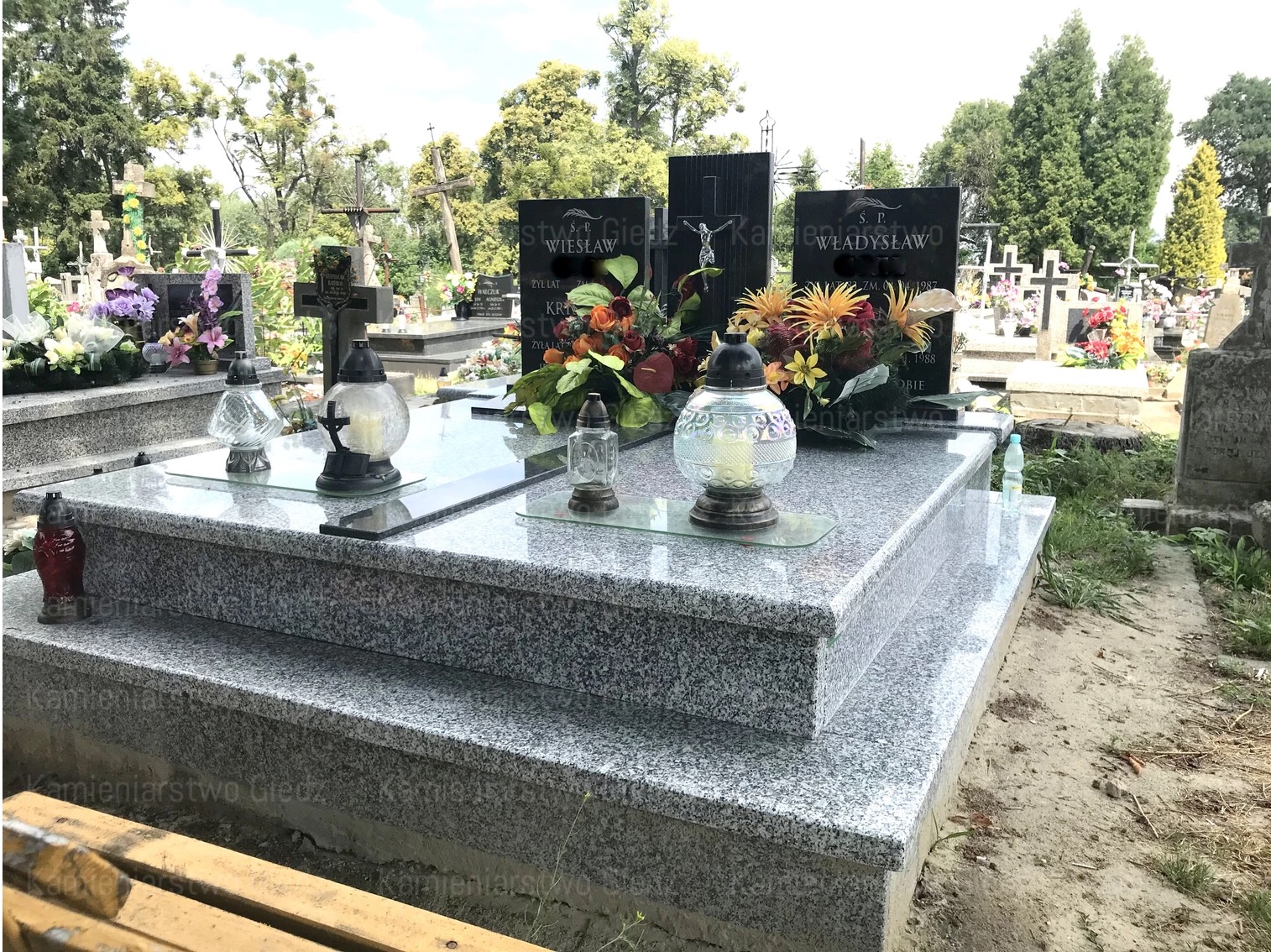 Granitowy pomnik nagrobny z dwoma płytami i szklanymi lampionami, ozdobiony kompozycjami kwiatowymi na cmentarzu.