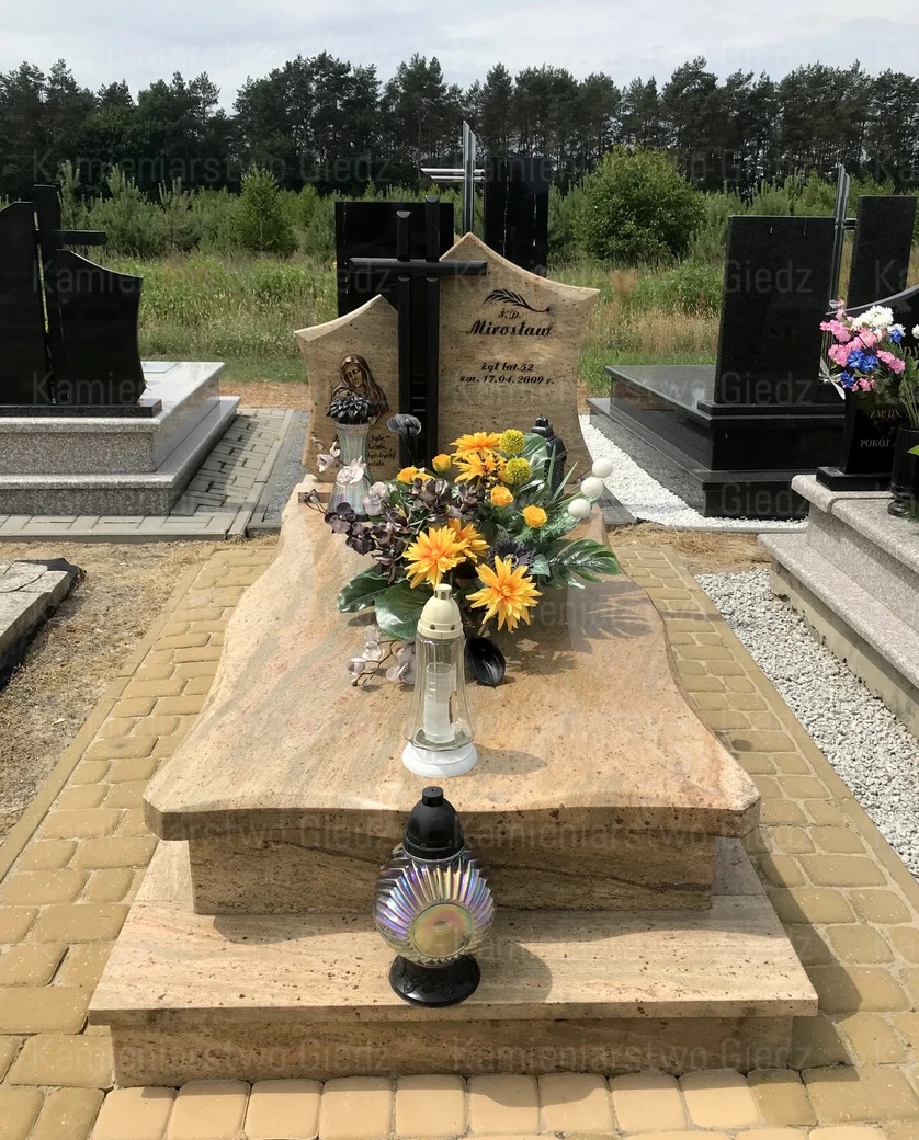 Jasny, granitowy nagrobek z wyrytym wizerunkiem kobiety i czarnym krzyżem, ozdobiony bukietem żółtych i fioletowych kwiatów oraz lampionami, otoczony kostką brukową i zielenią drzew.