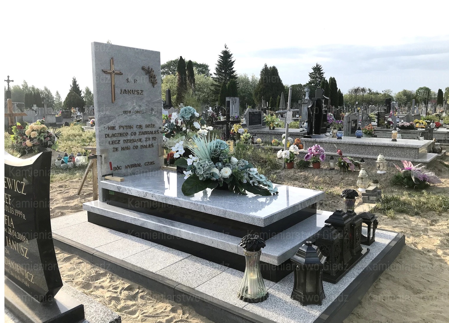 Jasnoszary, dwupoziomowy pomnik z inskrypcją i krzyżem, ozdobiony kompozycją kwiatową i lampionami, na tle cmentarza z innymi nagrobkami.