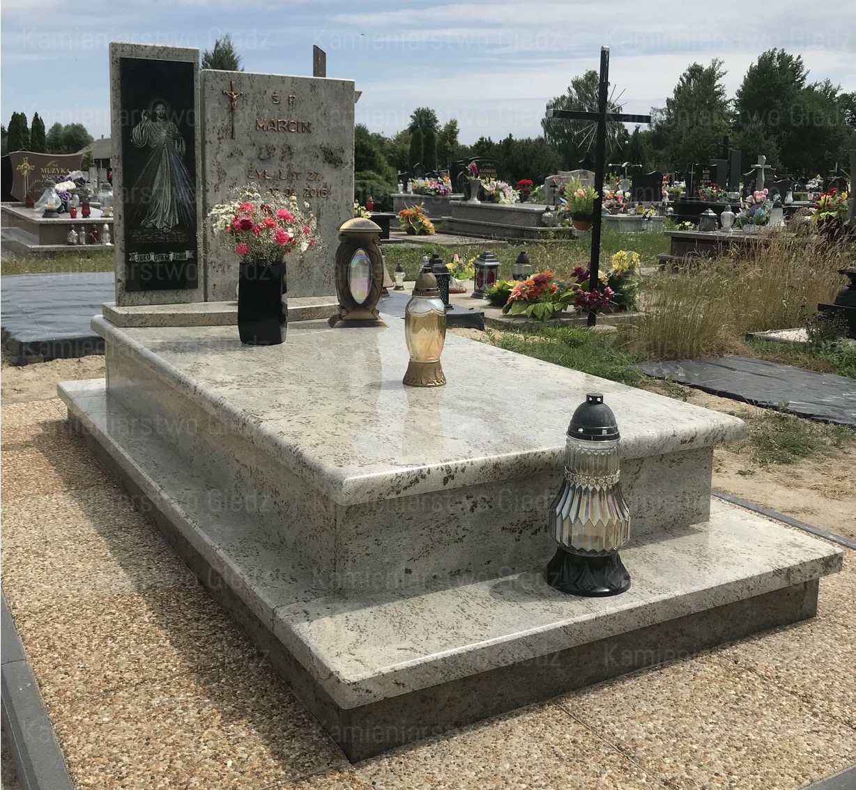 Jasny, dwupoziomowy pomnik z granitu z czarnym wazonem z kwiatami i lampionami na cmentarzu w słoneczny dzień.