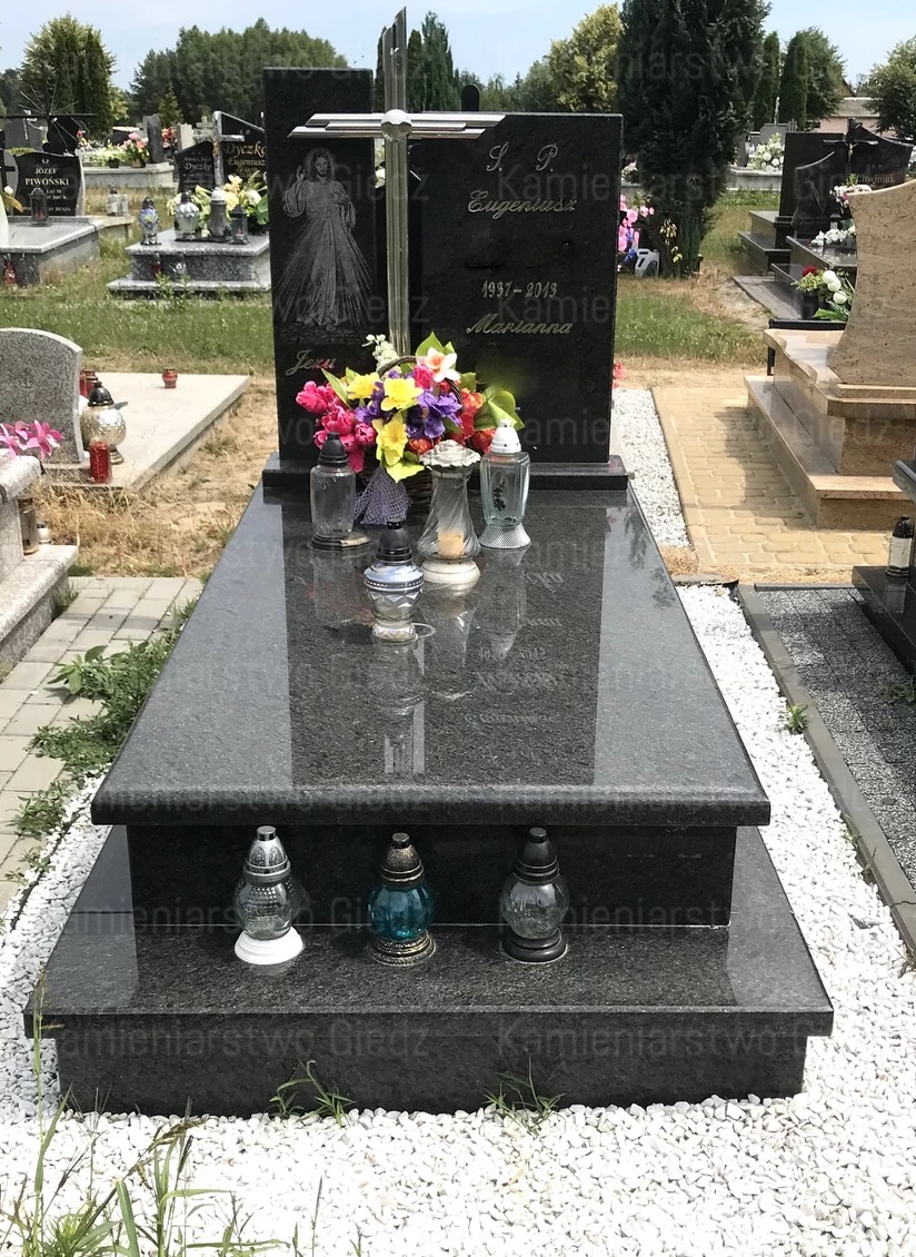 Dwupoziomowy, ciemny, granitowy pomnik z wygrawerowanym wizerunkiem postaci, ozdobiony kwiatami i lampionami, na tle cmentarza.