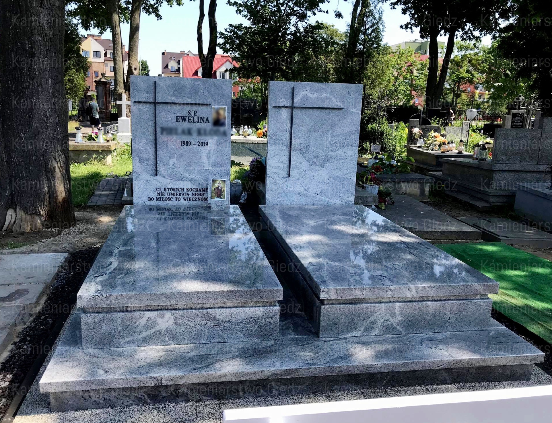 Dwa marmurowe nagrobki z czarnymi krzyżami i wygrawerowanymi inskrypcjami, otoczone zielenią cmentarza w słoneczny dzień.