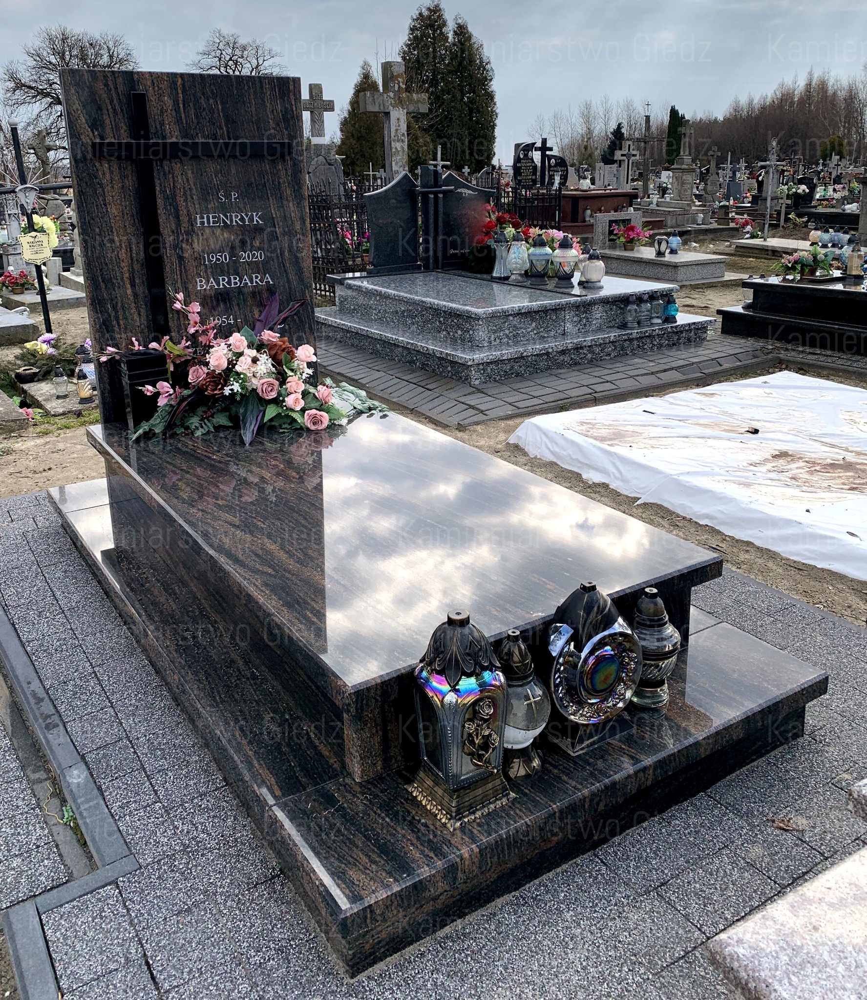 Dwupoziomowy, polerowany pomnik z ciemnego granitu z wyrytym krzyżem, kwiatami i zniczami na cmentarzu, odbijający pochmurne niebo.