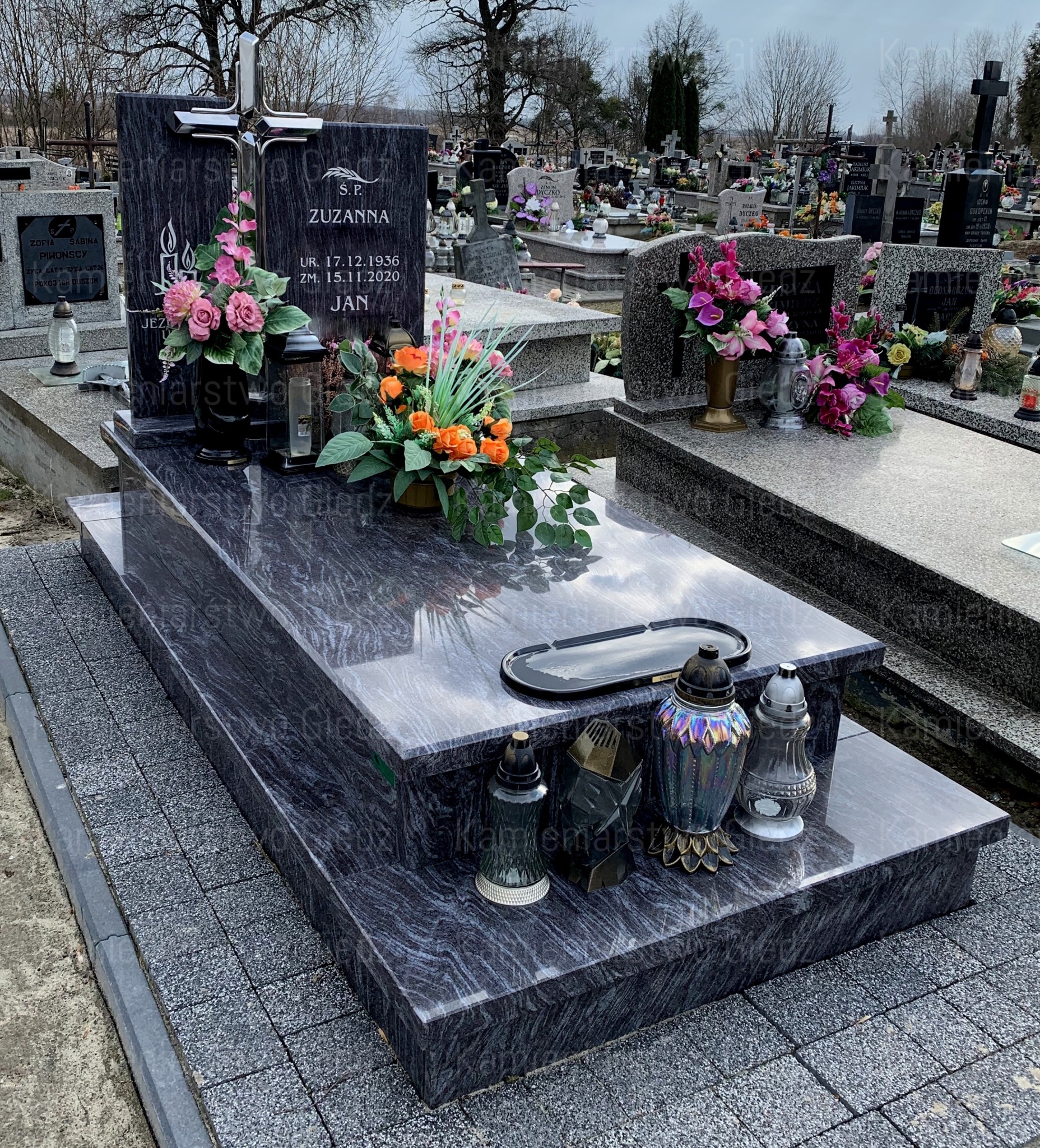 Granitowy nagrobek z czarną płytą inskrypcyjną, ozdobiony kwiatami i lampionami, na tle cmentarza.