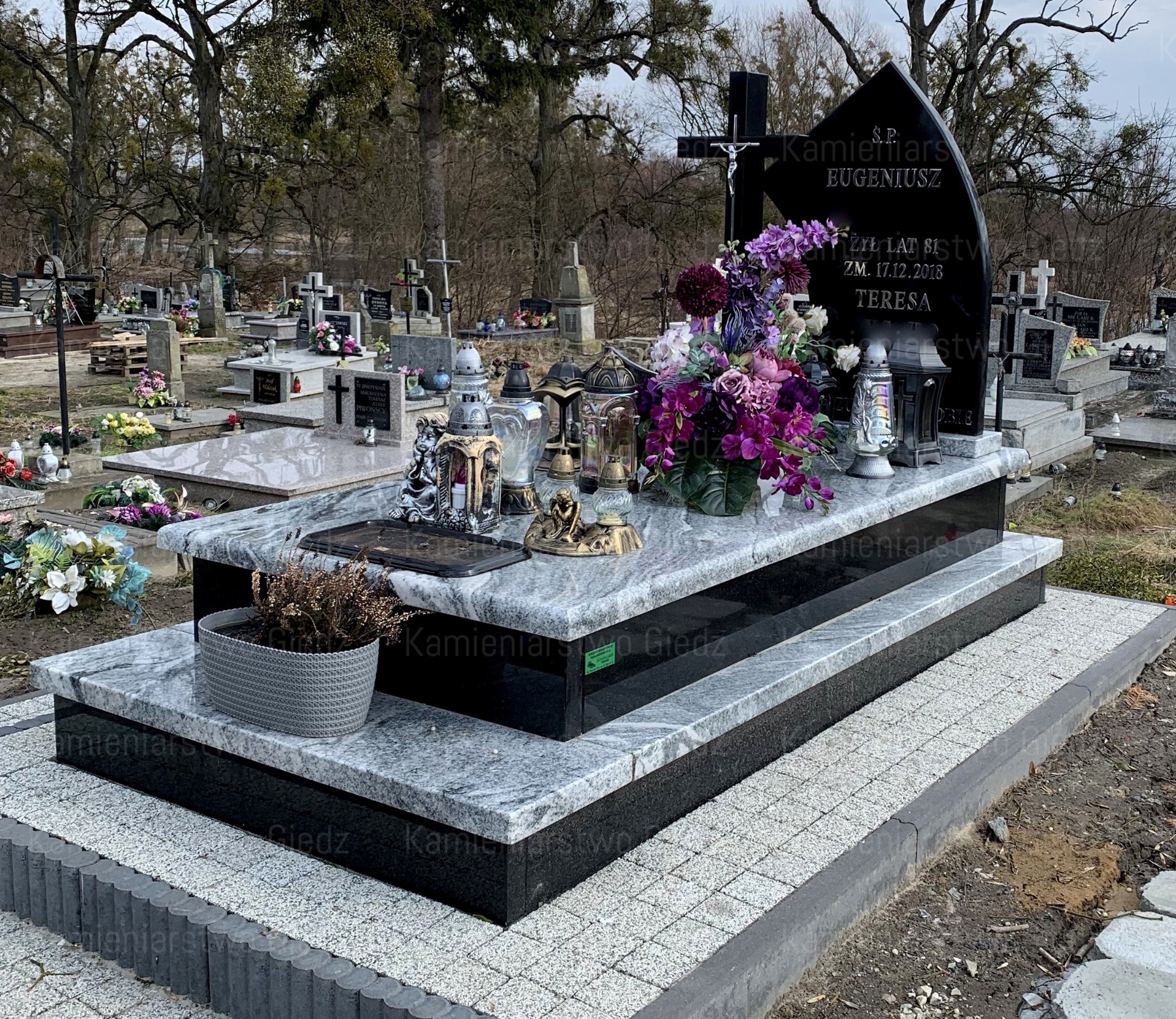 Dwupoziomowy pomnik z szarego granitu i czarnego kamienia, ozdobiony fioletowymi kwiatami, lampionami i figurkami aniołów, otoczony kostką brukową na cmentarzu.