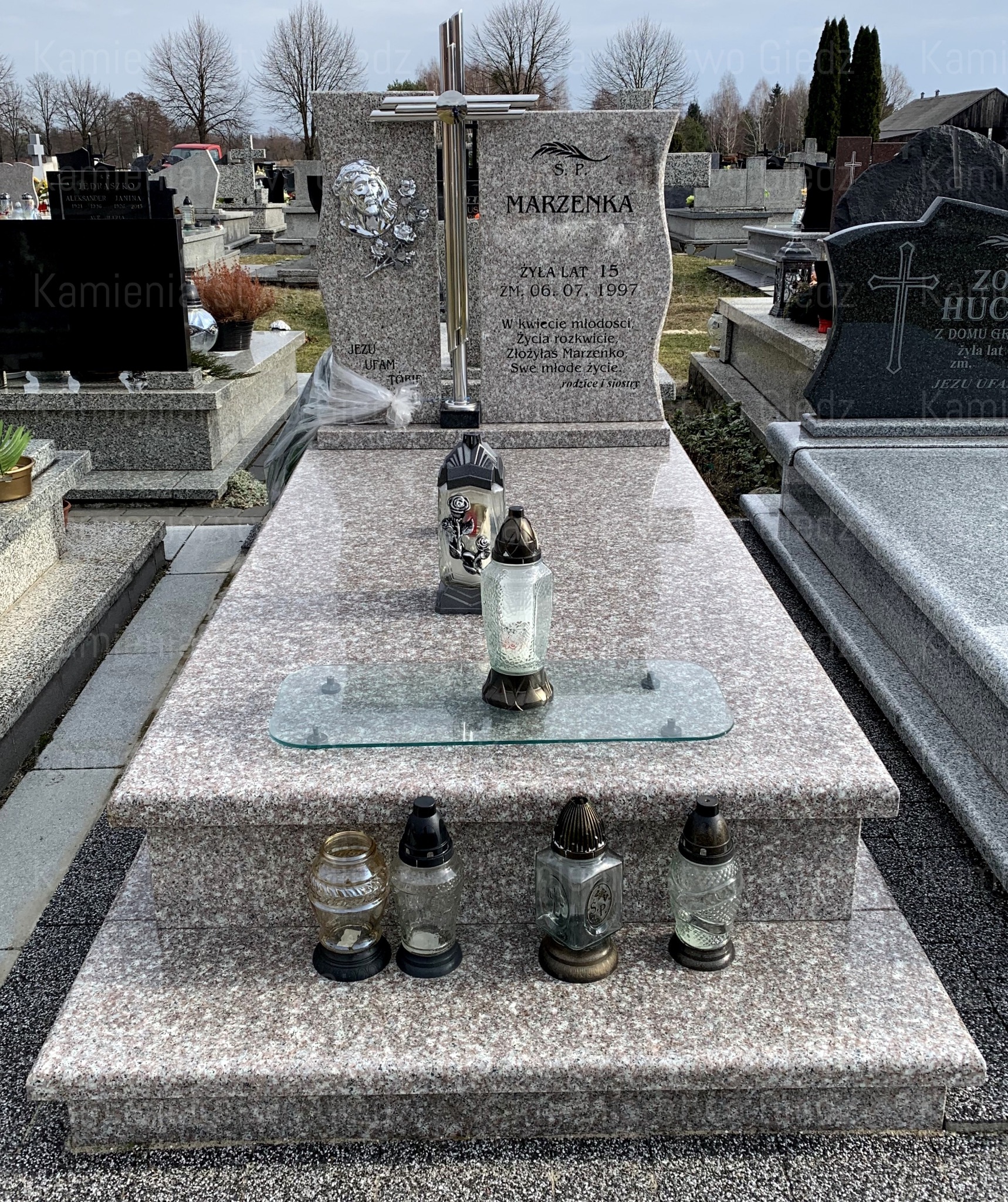 Granitowy pomnik nagrobny z metalowym krzyżem i wizerunkiem Jezusa, ozdobiony lampionami i szklaną półką na znicze, na tle innych nagrobków na cmentarzu.