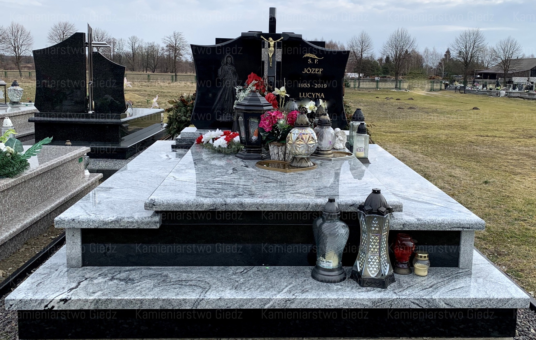 Dwupoziomowy, jasnoszary pomnik z czarnymi elementami, ozdobiony kwiatami i lampionami na cmentarzu w pochmurny dzień.