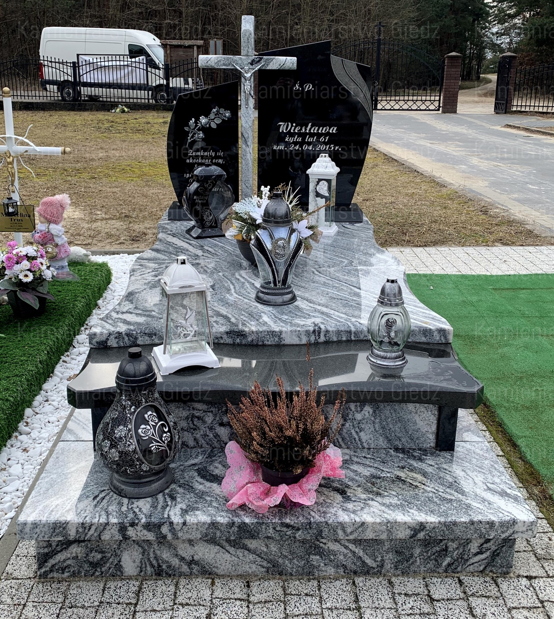 Granitowy pomnik z krzyżem, czarnymi tablicami i kwiatami, z widocznym nagrobkiem z jasnego, wzorzystego kamienia, ozdobiony lampionami i wrzosem w różowej osłonce, na tle cmentarza z alejką...