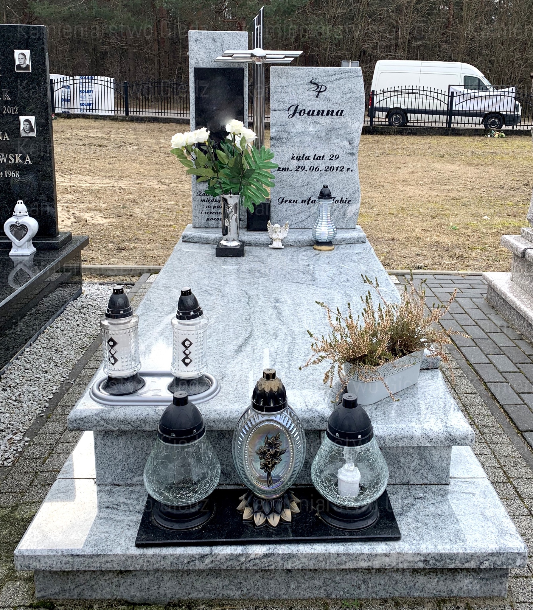 Granitowy pomnik z metalowym krzyżem, kwiatami w wazonie, figurką aniołka i zapalonymi lampionami na cmentarzu, z jasną kostką brukową w tle.