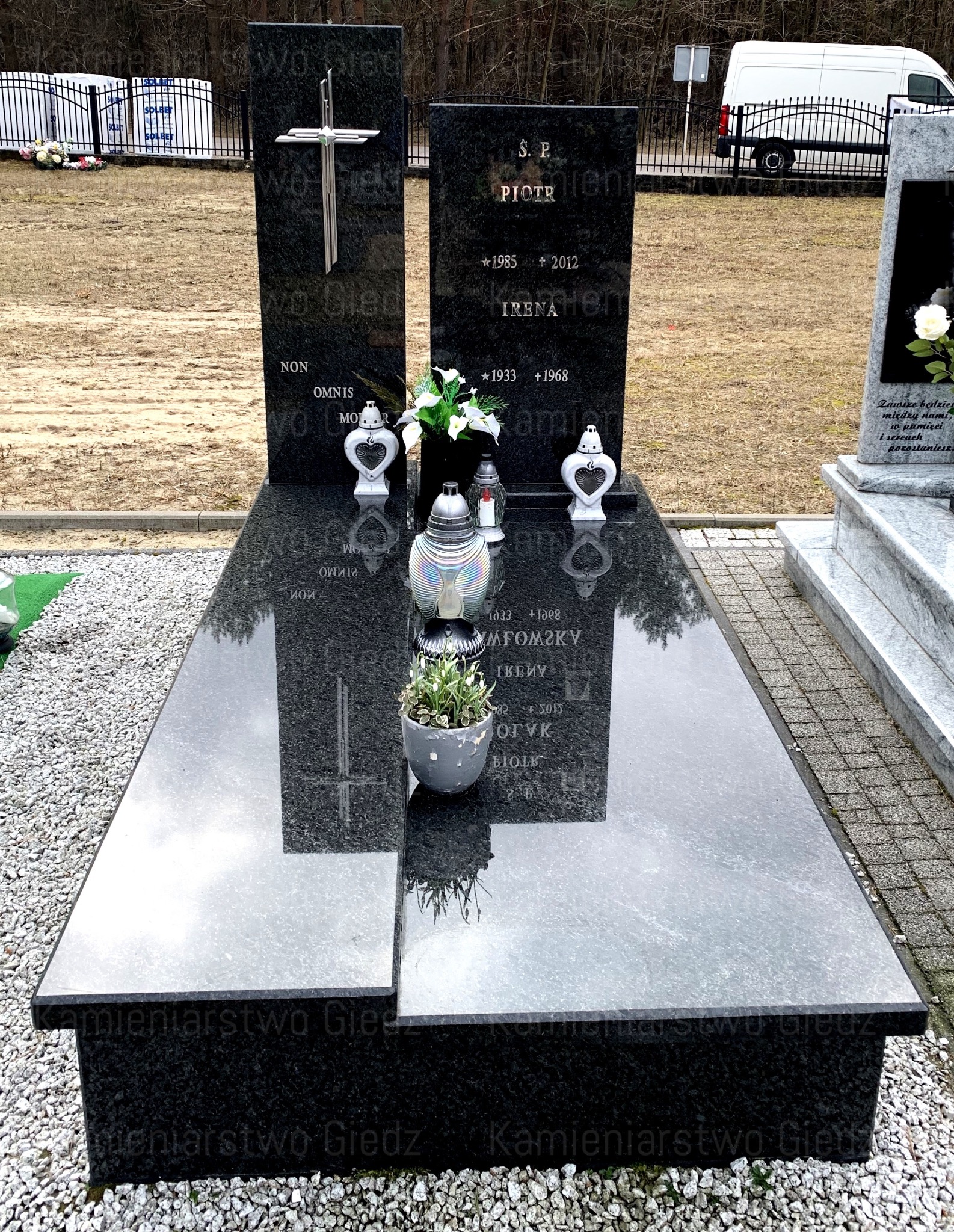 Czarny, granitowy pomnik z krzyżem i inskrypcjami, ozdobiony lampionami w kształcie serc, kwiatami i doniczką z rośliną, otoczony żwirem.