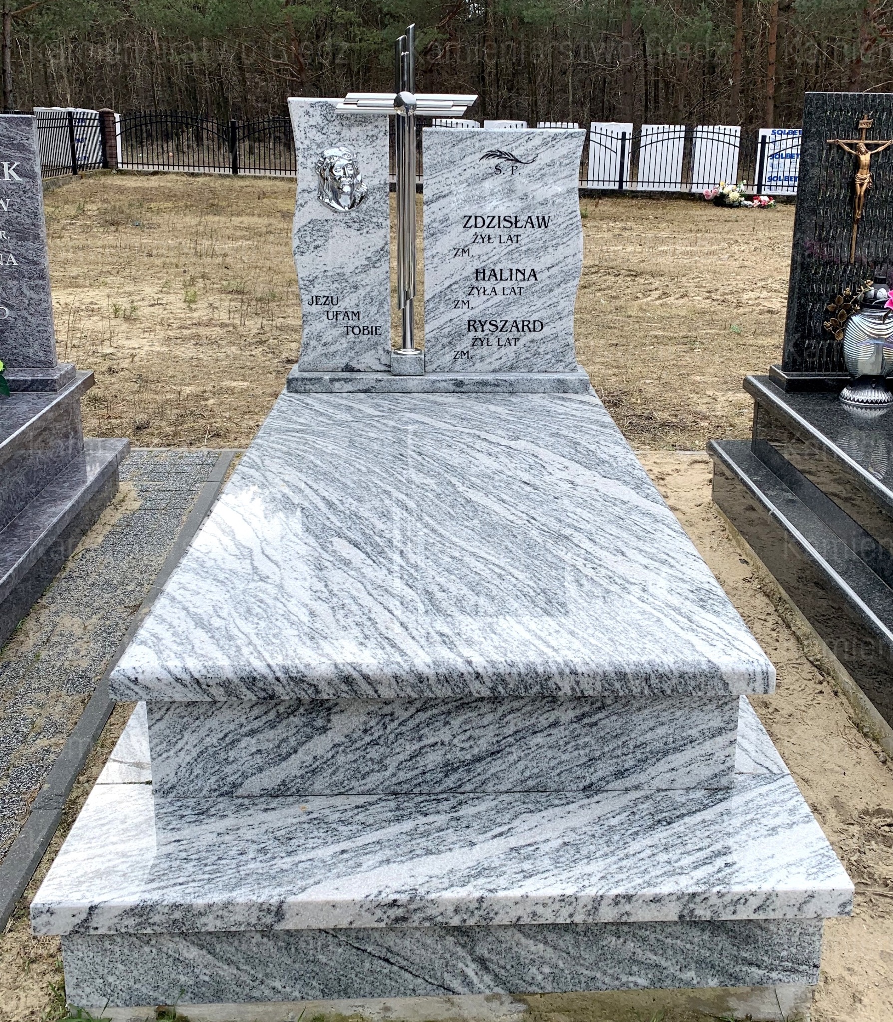 Granitowy nagrobek z jasnoszarego kamienia z wyraźnym, falistym wzorem, ozdobiony metalowym krzyżem i wizerunkiem Jezusa, na tle cmentarza i lasu.