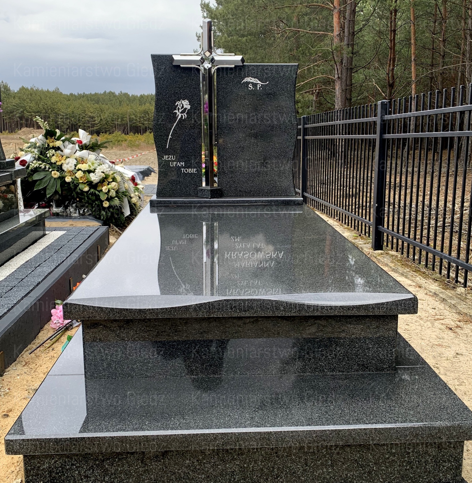 Czarny, błyszczący pomnik z krzyżem i wygrawerowanym napisem 'Jezu ufam Tobie' oraz motywem róży, z bukietem kwiatów obok, na tle drzew i metalowego ogrodzenia.