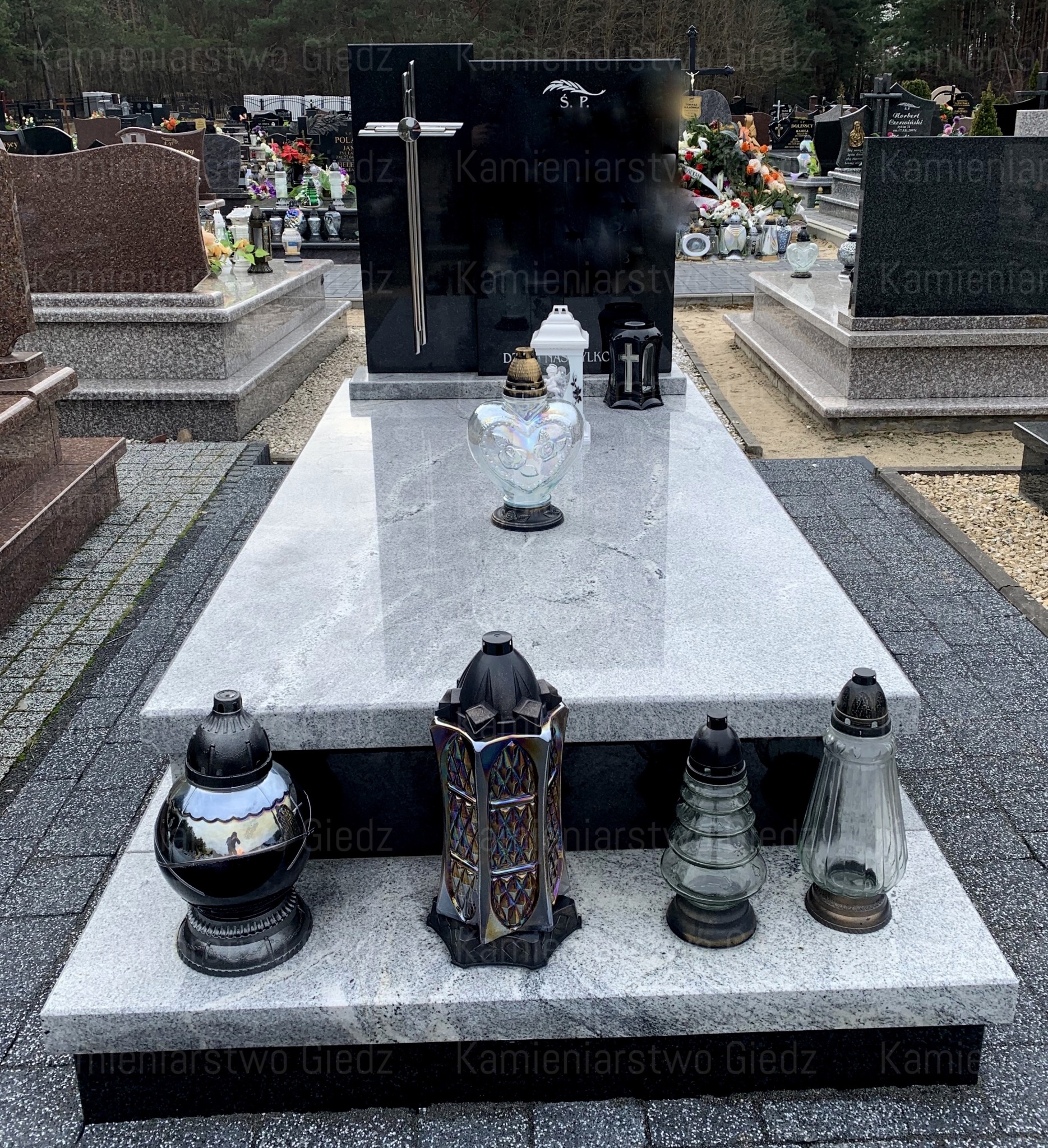 Czarny nagrobek z jasnoszarym blatem, ozdobiony metalowym krzyżem i różnorodnymi lampionami na cmentarzu.
