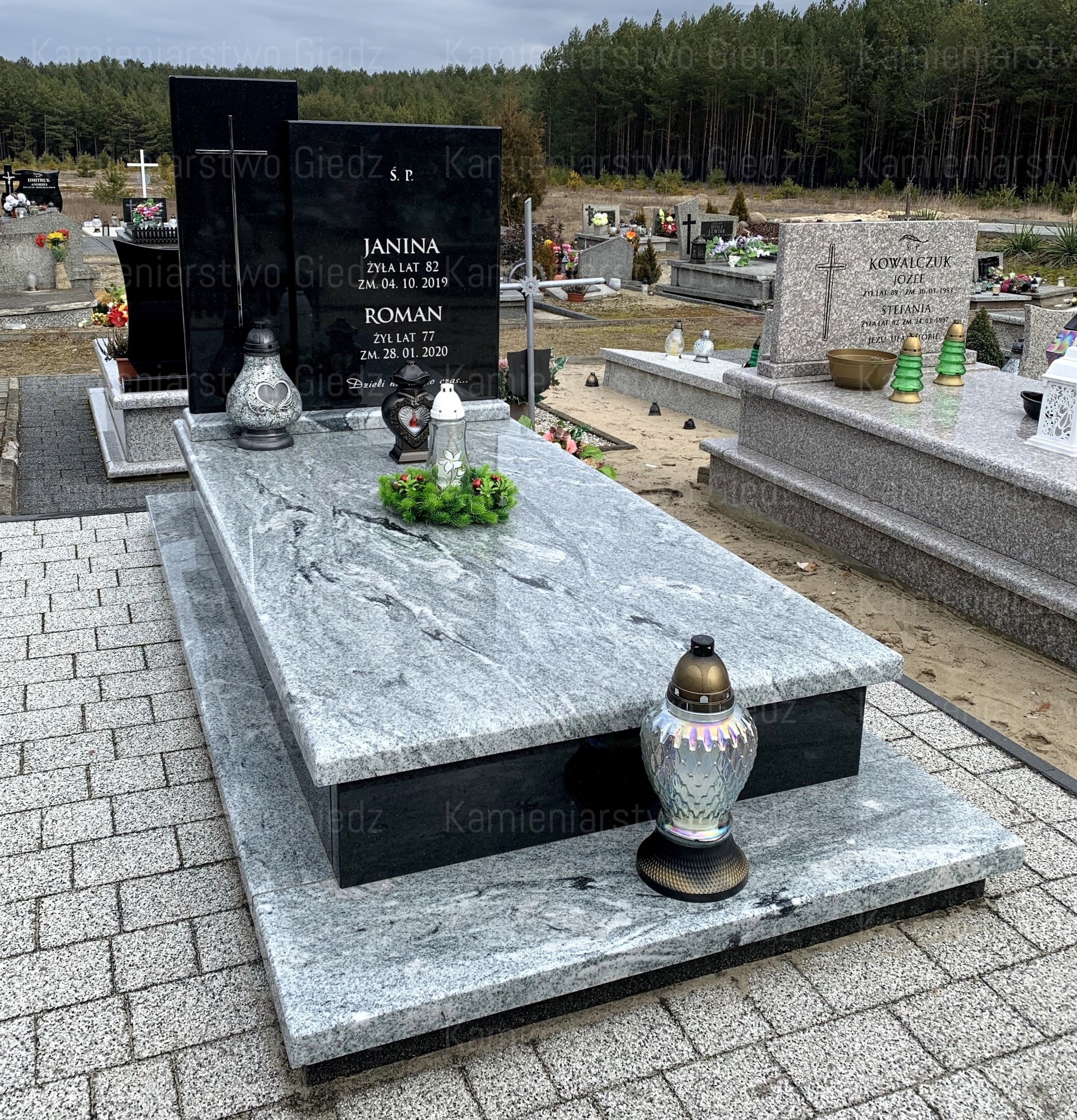 Granitowy pomnik z czarną tablicą nagrobną, ozdobiony lampionami i wieńcem, na cmentarzu z widocznym lasem w tle.