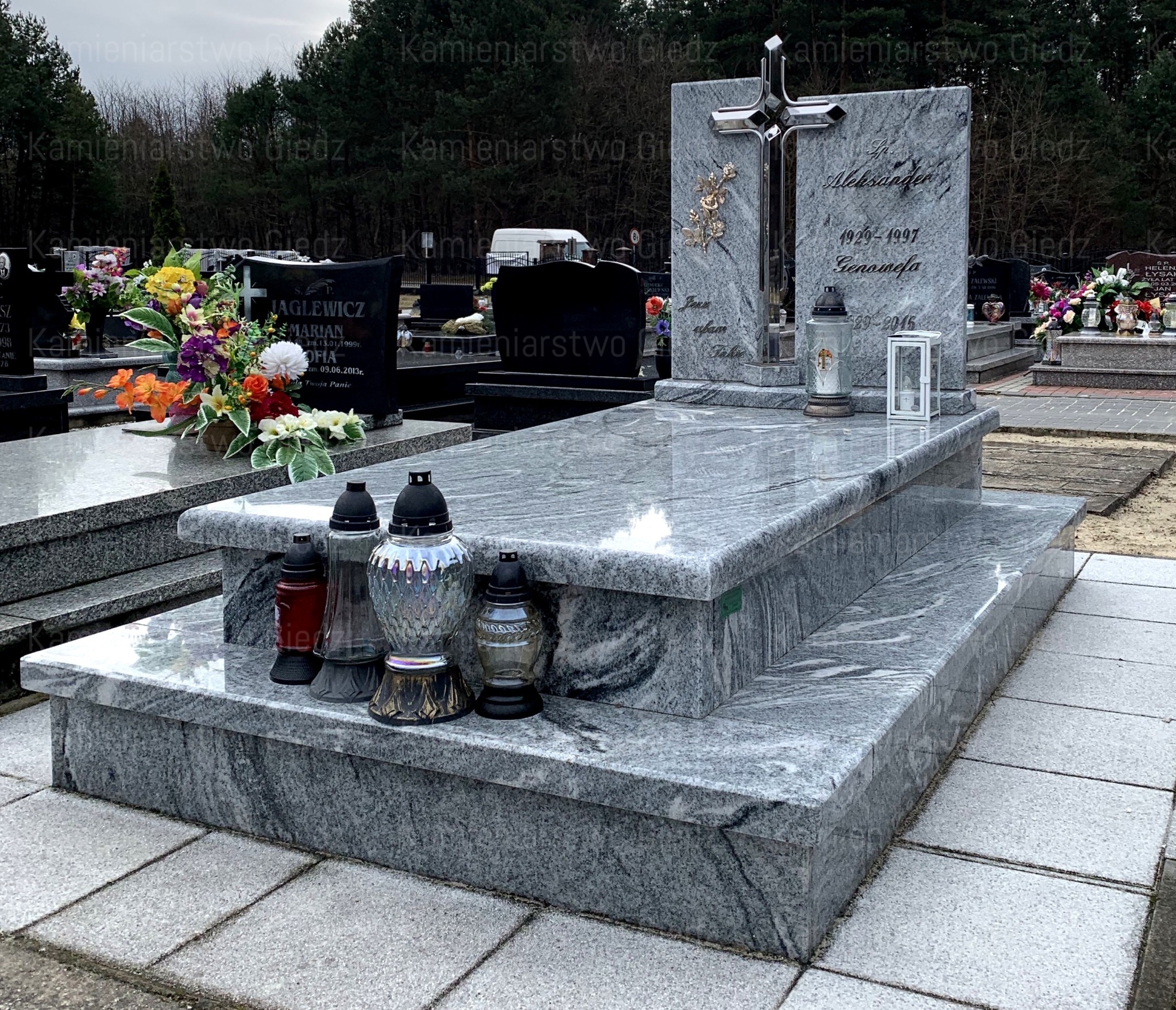 Dwupoziomowy pomnik z szarego kamienia z krzyżem ze stali nierdzewnej i metalową aplikacją kwiatową, otoczony zniczami i innymi nagrobkami na cmentarzu.