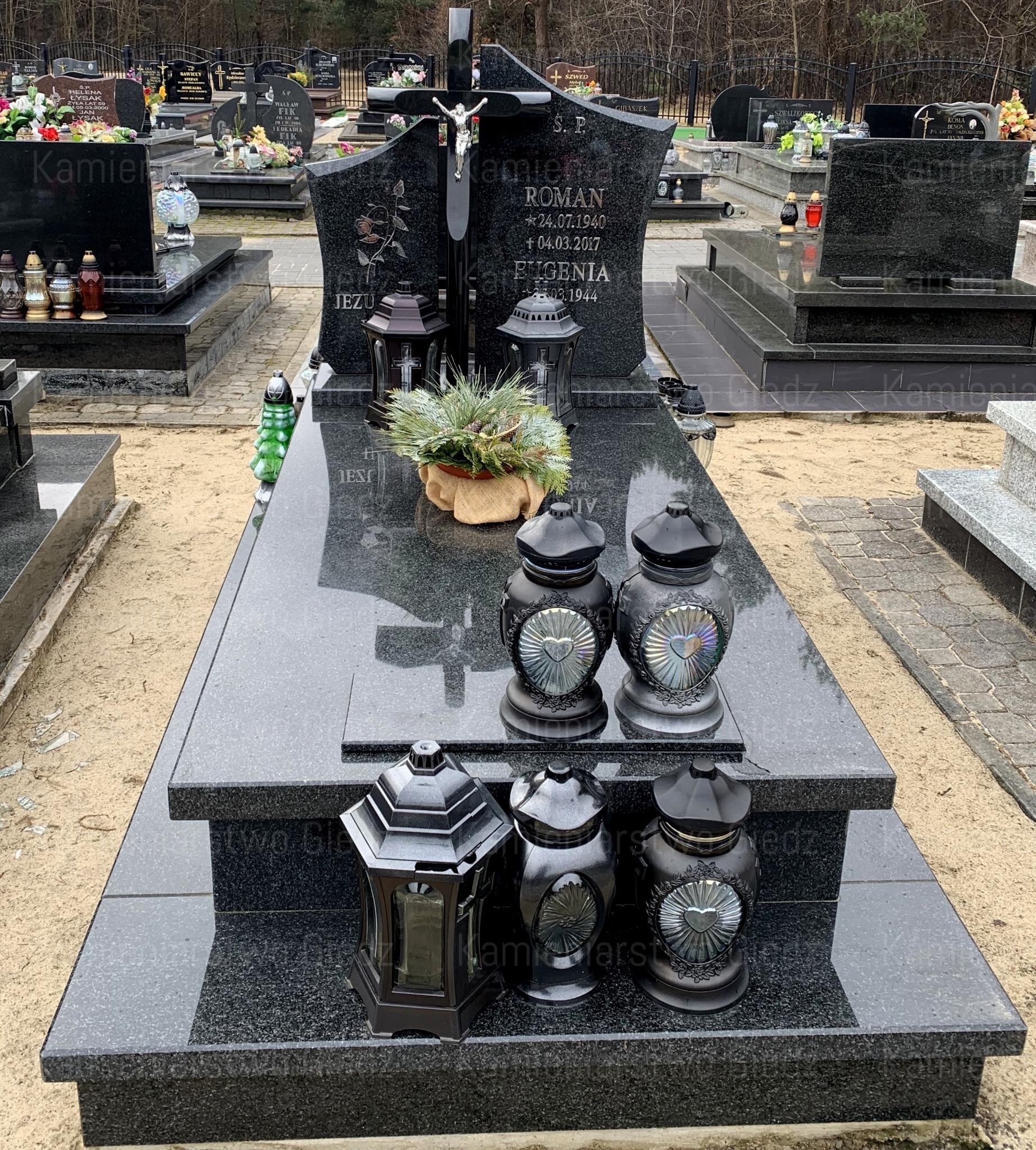 Czarny, granitowy pomnik z krzyżem i inskrypcjami na cmentarzu w Świerżach, ozdobiony lampionami i stroikiem. Widoczne inne nagrobki w tle.