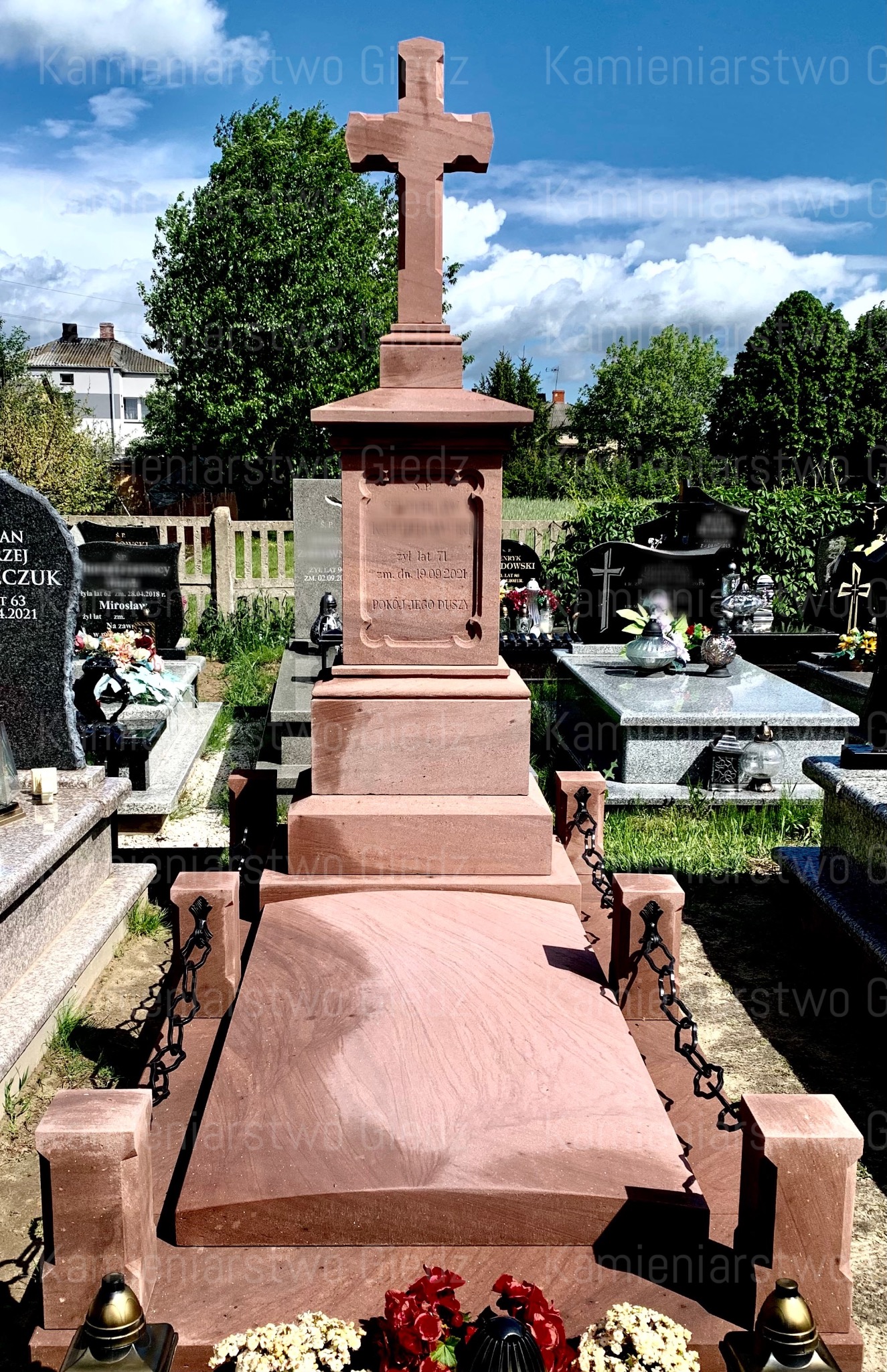 Czerwony piaskowiec: nagrobek z krzyżem na cmentarzu, otoczony łańcuchem, z widocznymi kwiatami i zniczami.