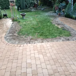 Świeżo ułożona, wilgotna ścieżka z kostki brukowej w ogrodzie, otaczająca trawnik przygotowany do nasadzeń, widoczny wąż ogrodowy i roślinność w tle.