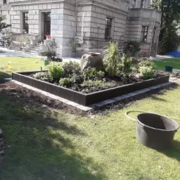 Ogródek warzywny wzniesiony na trawniku, obudowany ciemnymi, prostokątnymi elementami, z kamieniem centralnym i różnorodnymi roślinami, na tle klasycznej architektury budynku.