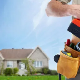 Mężczyzna z pasem narzędziowym trzyma wiertarkę, w tle klucz w kształcie domu i rozmyty widok domu jednorodzinnego na tle błękitnego nieba.