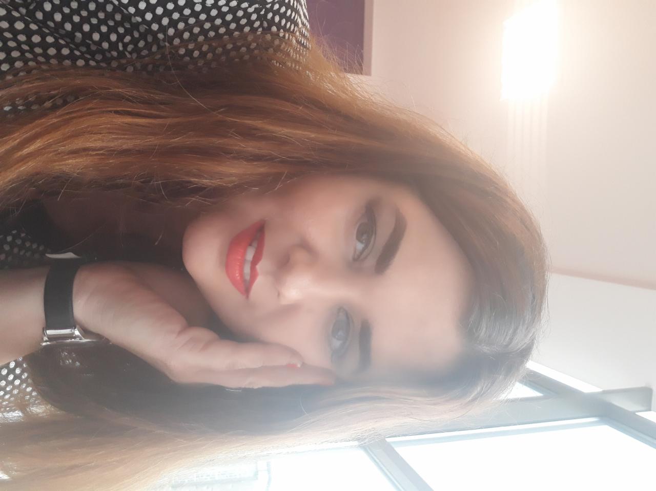 Portret kobiety z czerwonymi ustami, opierającej głowę na dłoni z widocznym zegarkiem na tle okna i wzorzystej bluzki.