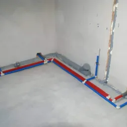 Wnętrze w trakcie instalacji hydraulicznej: rury w kolorze czerwonym i niebieskim prowadzone po podłodze i wzdłuż ścian, szare rury kanalizacyjne, widoczne przyłącza do grzejników i armatury.