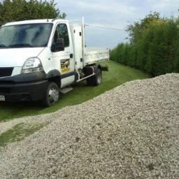 Biała wywrotka Renault zaparkowana na trawie obok sporej pryzmy jasnych kamieni, z żywopłotem w tle.