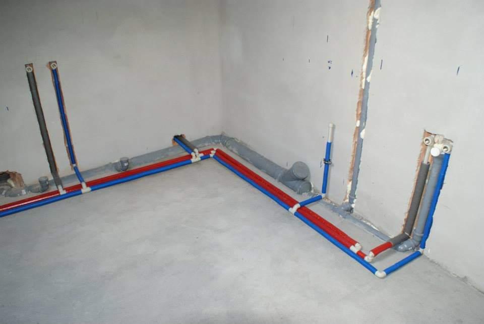 Wnętrze w trakcie instalacji hydraulicznej: rury w kolorze czerwonym i niebieskim prowadzone po podłodze i wzdłuż ścian, szare rury kanalizacyjne, widoczne przyłącza do grzejników i armatury.