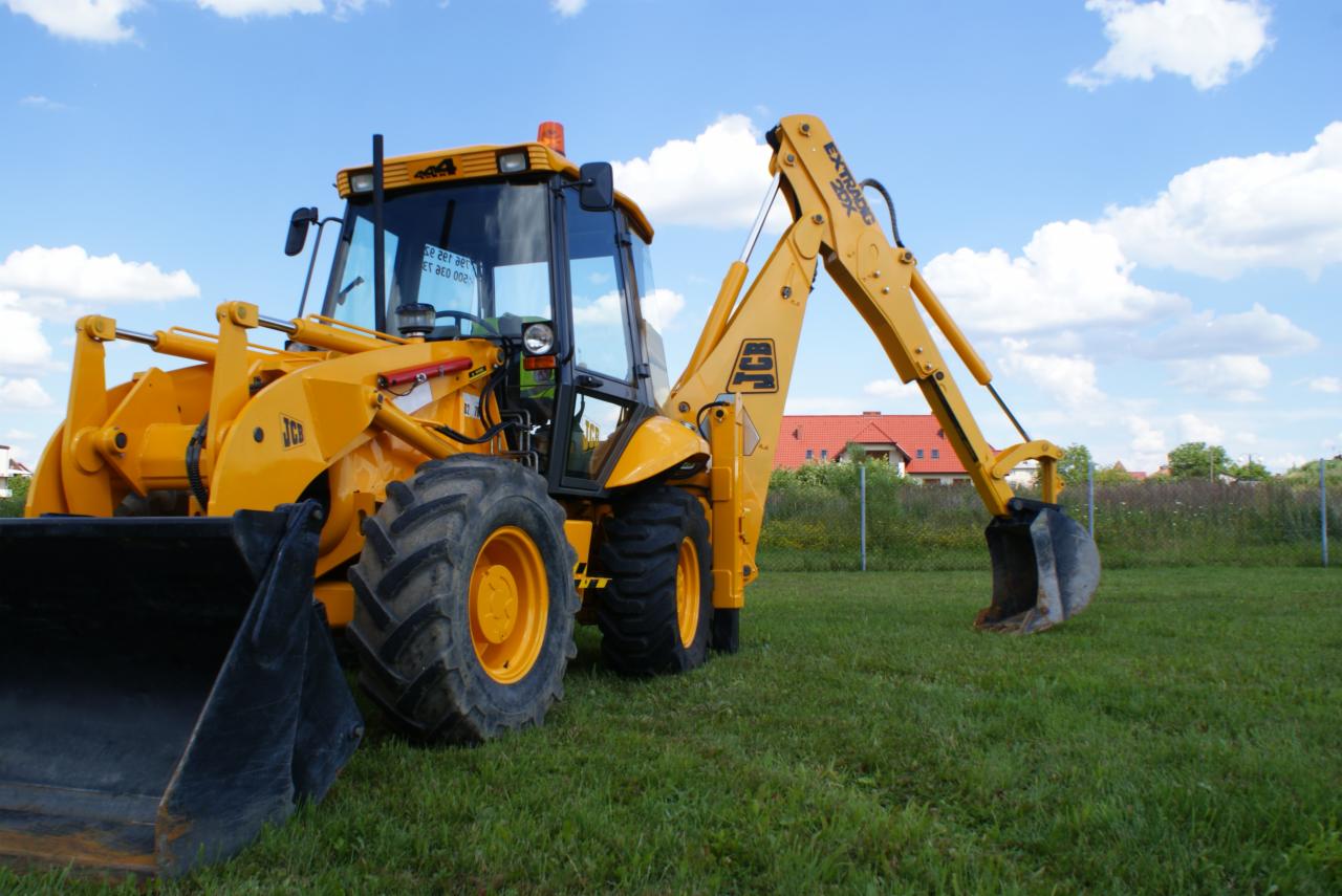 Żółta koparko-ładowarka JCB na trawiastym terenie, widoczny łyżka z przodu i ramię koparki z tyłu, w tle dom z czerwoną dachówką i błękitne niebo z chmurami.