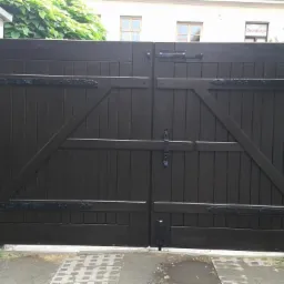 Dwuskrzydłowa, ciemnobrązowa brama wjazdowa z dekoracyjnymi zawiasami, zamontowana między białymi słupkami w Bochni.