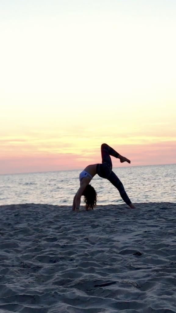 Kobieta w stroju sportowym wykonuje mostek z jedną nogą uniesioną na piaszczystej plaży o zachodzie słońca.