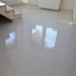 Błyszcząca, jasna podłoga wyłożona kwadratowymi płytkami ceramicznymi, odbijająca światło okna, widoczne schody w surowym stanie i grzejnik na ścianie.